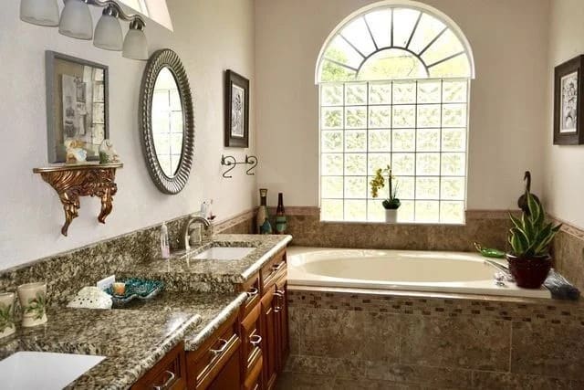 A bright bathroom with a granite countertop, wooden cabinets, a large mirror, a built-in bathtub, potted plants, and a large arched window letting in natural light. Framed art is on the walls.