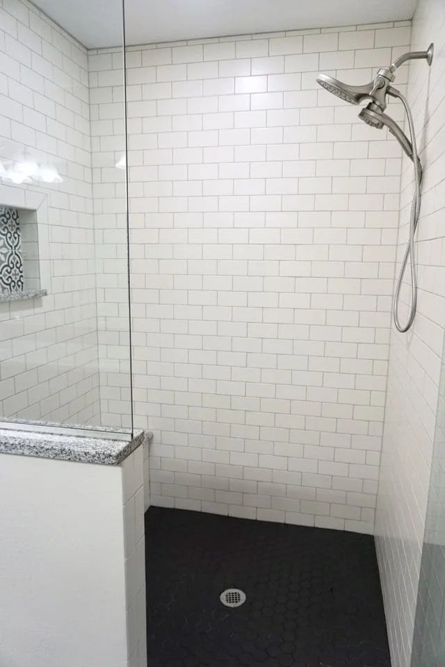 A modern shower with white subway tile walls, a built-in shelf with patterned tiles, a silver showerhead, a frameless glass divider, and a dark hexagon-tile floor with a central drain.