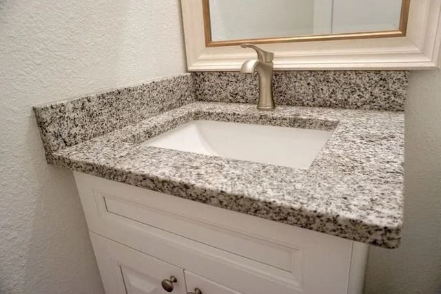 A bathroom vanity with a rectangular, undermount sink and a granite countertop, set in a corner. The vanity is white with silver handles, and there is a silver faucet and a framed mirror above.