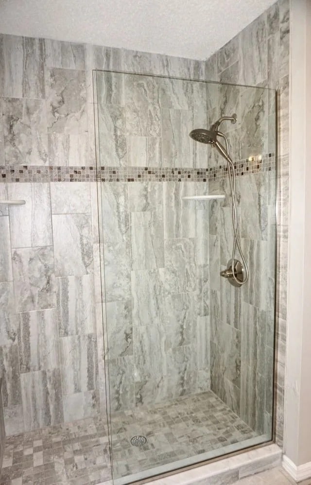 A modern walk-in shower with gray marble-patterned tiles, a glass door, a rainfall showerhead, a handheld shower attachment, and two small white corner shelves.
