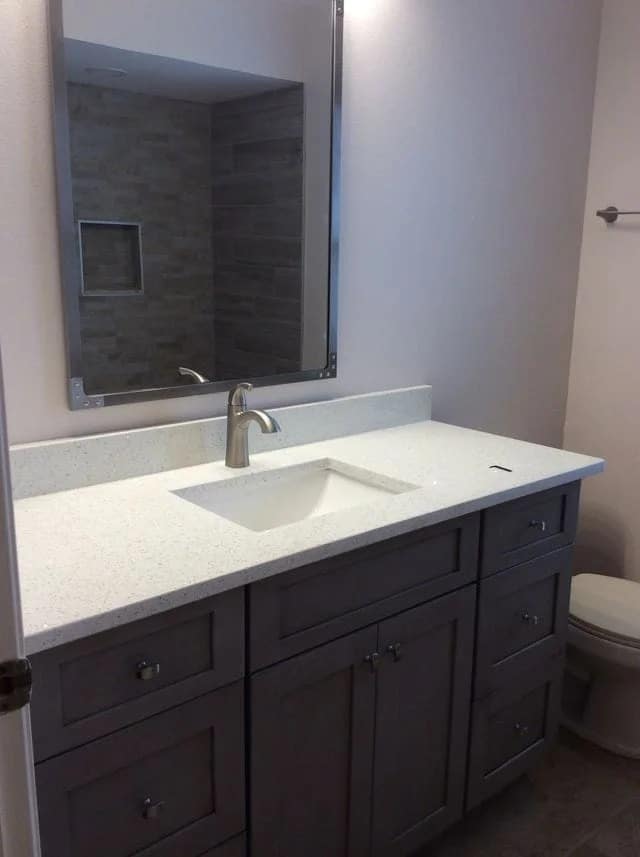 A modern bathroom with a gray vanity, white countertop, under-mount sink, silver faucet, large wall mirror, and a toilet to the right. The background shows a tiled shower area.