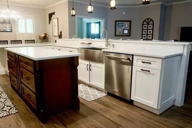 Modern kitchen with white cabinets, a stainless steel farmhouse sink, dishwasher, dark wood island, pendant lights, and hardwood floors. A patterned rug and wall art decorate the space.