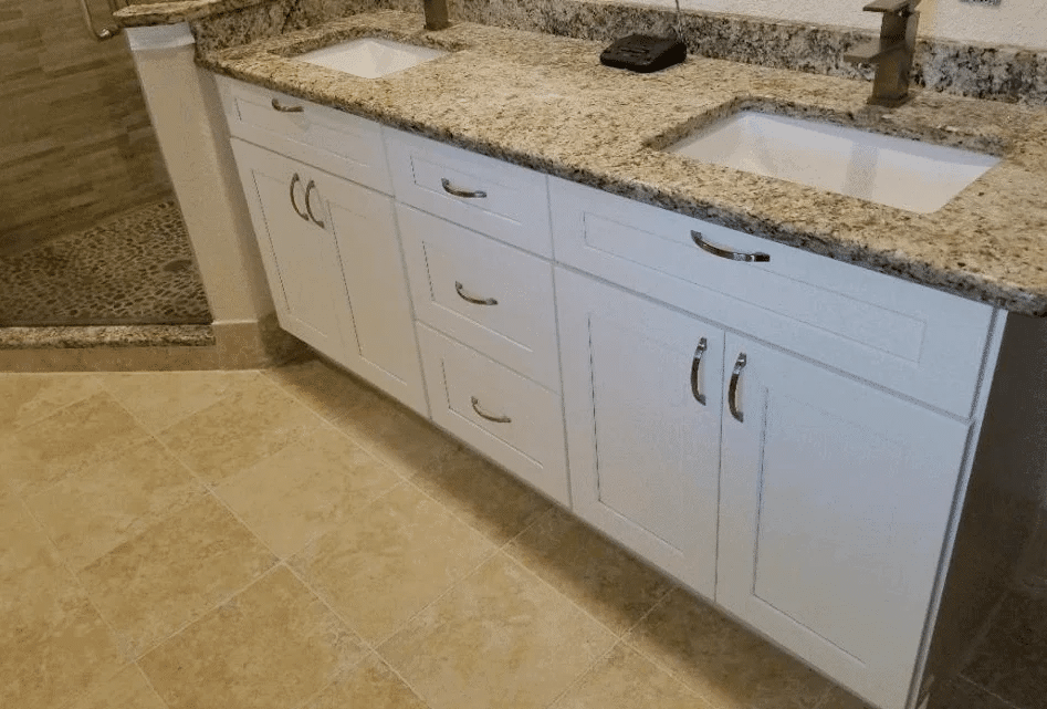A bathroom with a granite countertop, two rectangular sinks, white cabinets and drawers, and beige tile flooring. A walk-in shower with stone flooring is visible to the left.