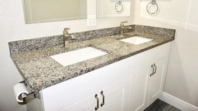 A bathroom vanity with a speckled granite countertop, two rectangular sinks, modern faucets, white cabinets, and a toilet paper holder mounted on the left side.