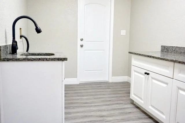 A modern laundry room with white cabinets, granite countertops, a black faucet, light gray walls, and wood-style flooring. A closed white door is at the end of the room.