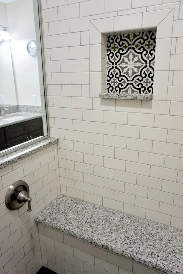 A shower with white subway tiles, a built-in granite bench, a silver faucet, and a recessed shelf featuring black and white decorative tiles as an accent. A mirror and countertop are visible in the background.