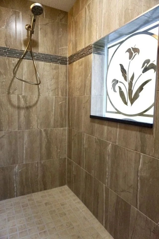A tiled walk-in shower with beige marble walls, a mosaic accent strip, a wall-mounted showerhead, and a window featuring frosted glass with a floral design.