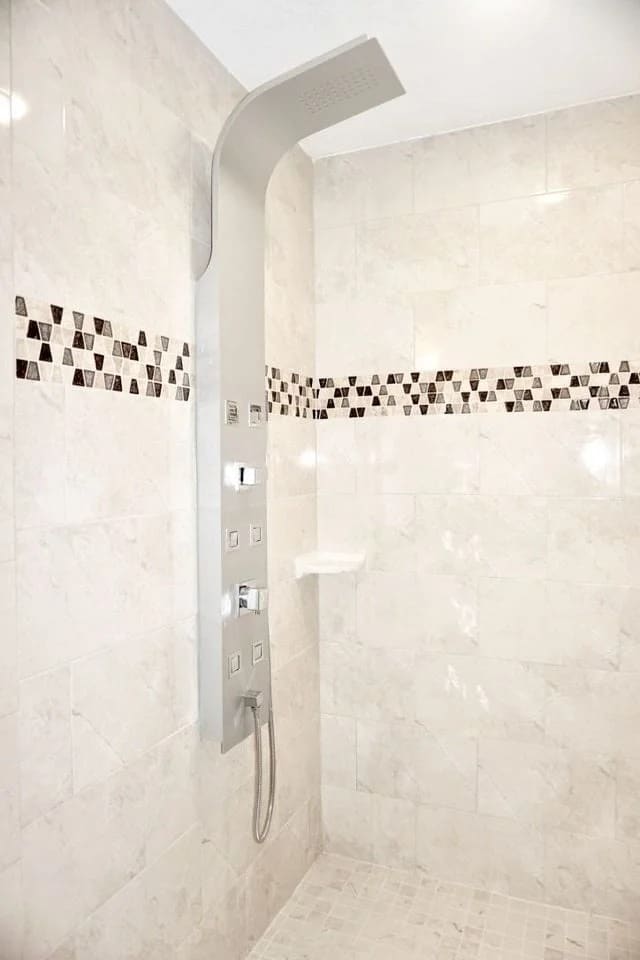 A modern shower with beige marble tiles, a row of decorative mosaic tiles, and a sleek silver shower panel with multiple controls and a handheld showerhead.