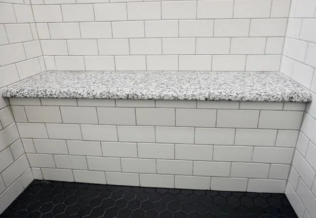 A shower corner with white subway tile walls, a speckled granite bench, and a floor of black hexagonal tiles.