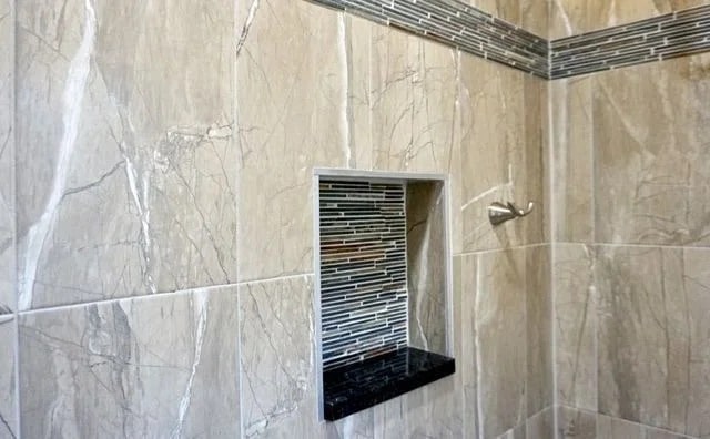 Shower wall with beige marble tiles, a recessed shelf lined with small horizontal black, gray, and white tiles, and a matching horizontal accent tile strip along the upper part of the wall.