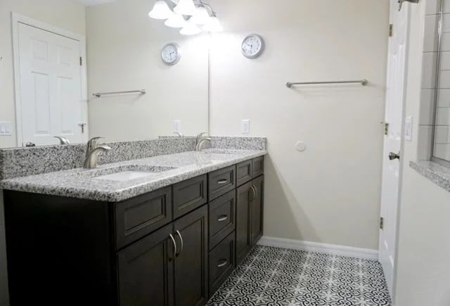 A modern bathroom with double sinks, dark wood cabinets, a granite countertop, a large mirror, two wall clocks, towel racks, and patterned tile flooring.