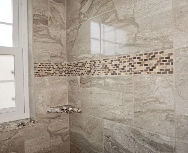 A tiled shower wall with large beige and gray marble tiles, a horizontal mosaic tile accent strip, a small corner shelf, and a window on the left side.