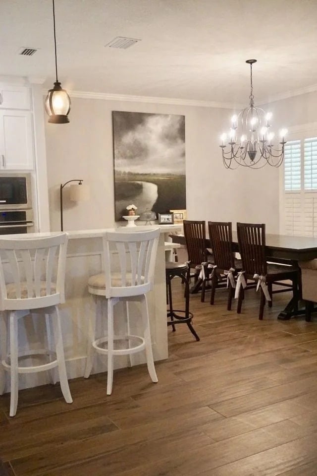 A cozy dining area with a wooden floor, white breakfast bar with two white chairs, dark wooden dining table and matching chairs, a modern chandelier, and a large landscape painting on the wall.