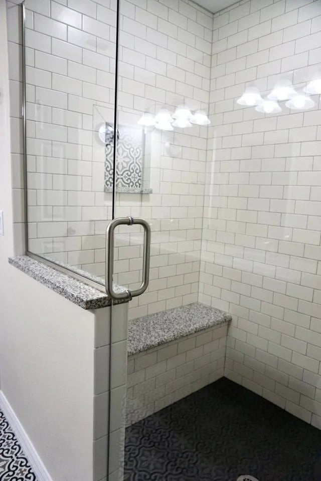 A modern walk-in shower with white subway tile walls, a glass door with a metal handle, a built-in granite bench, and a dark patterned tile floor. Bright lights are reflected on the glass.