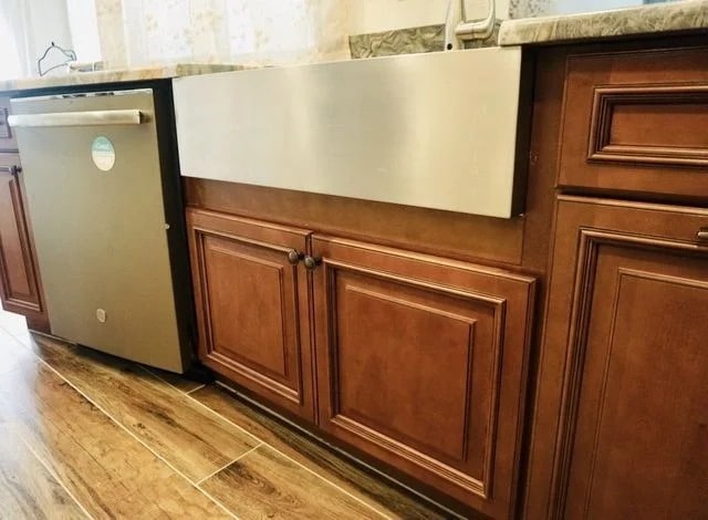 A modern kitchen with brown wooden cabinets, a stainless steel farmhouse sink, a built-in dishwasher, and wood-look tile flooring. The countertop is light-colored with a marble pattern.