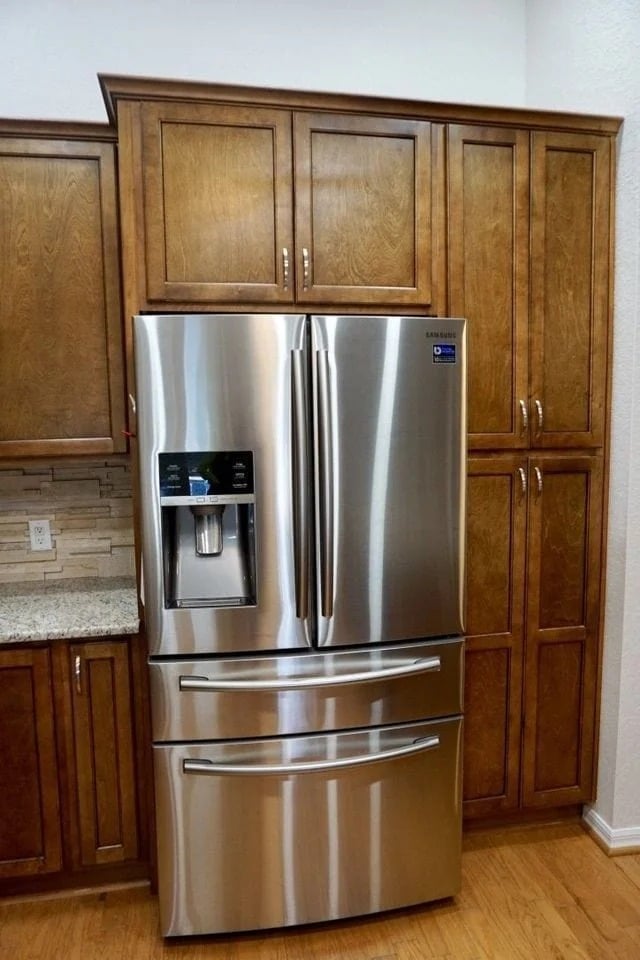 A stainless steel French door refrigerator with two freezer drawers is placed in a kitchen, partially blocking two tall wooden cabinets above and to the side. The fridge features a water and ice dispenser.