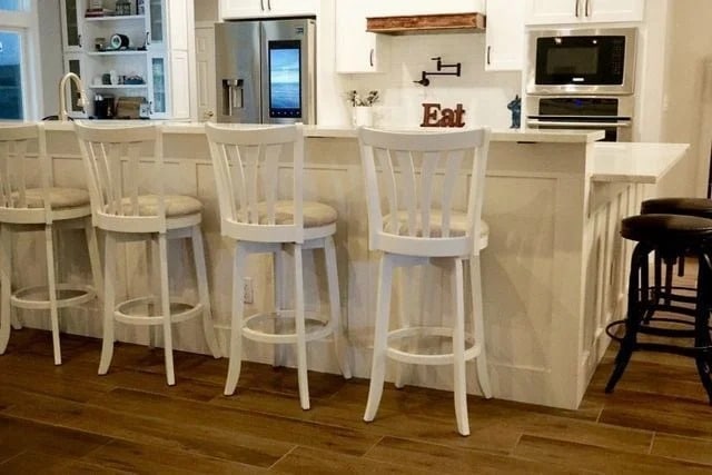 A kitchen with a white island, four white barstools with cushioned seats, two dark barstools, and wooden flooring. The background shows a stove, oven, microwave, fridge, and a sign that says "Eat.