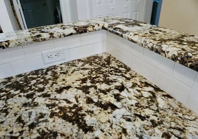 A kitchen countertop with a brown, black, and white granite pattern, white tile backsplash, and an electrical outlet on the wall. The counter has multiple levels and clean, modern finishes.