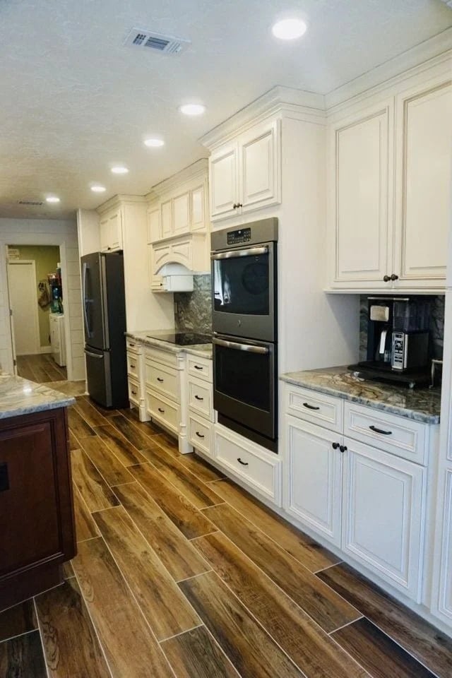 Modern kitchen with white cabinets, stainless steel appliances, double ovens, marble countertops, wood-look tile flooring, and recessed ceiling lights. A coffee maker sits on the counter to the right.