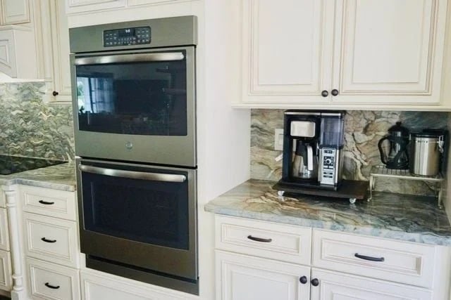 A modern kitchen with a built-in double oven on the left and a coffee maker with a grinder on a marble countertop to the right; white cabinets and drawers are visible throughout.