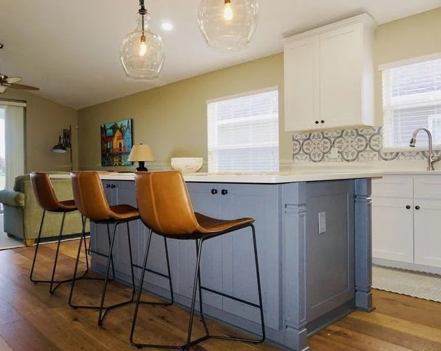 A modern kitchen with a blue island, three brown leather barstools, pendant lights, wood flooring, white cabinets, patterned backsplash, and large windows letting in natural light.
