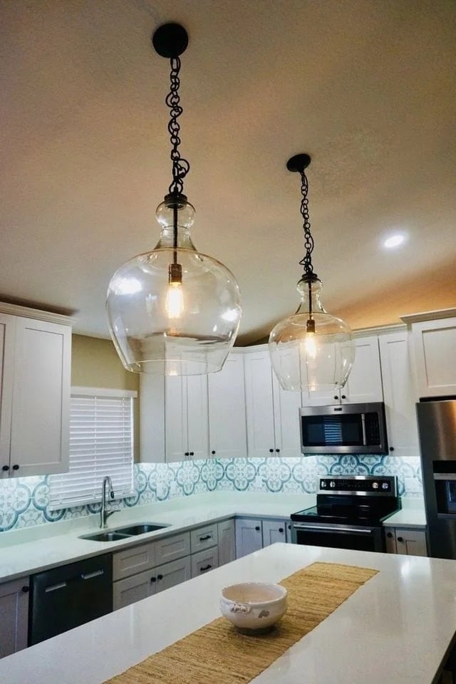 Modern kitchen with white cabinets, patterned blue and white backsplash, stainless steel appliances, a white island with a tan runner, and two large glass pendant lights hanging from the ceiling.