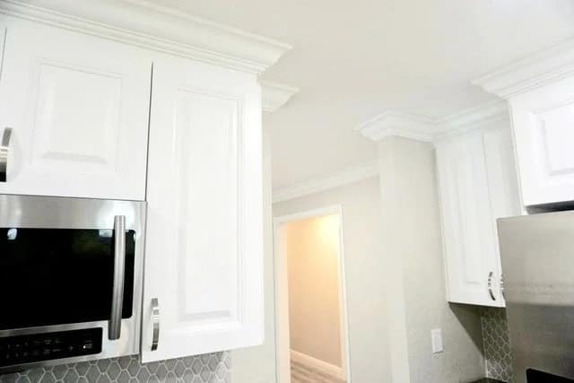 Bright kitchen with white cabinets, stainless steel appliances, and a geometric tiled backsplash. An open doorway leads to a hallway with neutral-colored walls and crown molding along the ceiling.