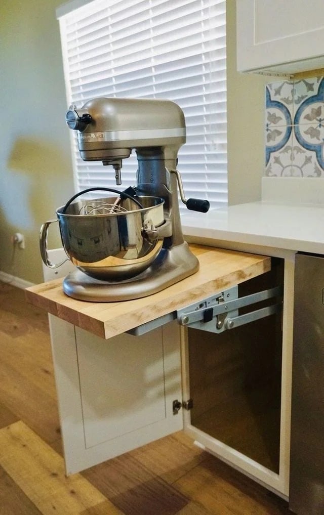 A stand mixer sits on a wooden pull-out shelf extended from a kitchen cabinet, with a window and tiled backsplash in the background.