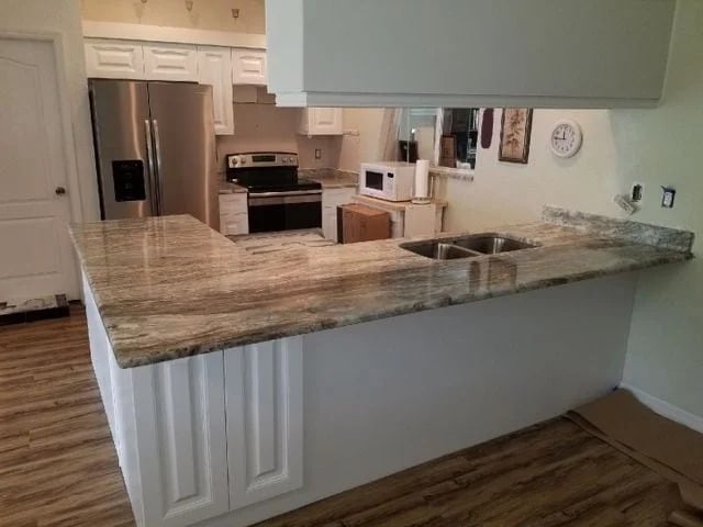 A modern kitchen with a U-shaped granite countertop, stainless steel appliances, white cabinets, a double sink, microwave, and wood-patterned flooring. A wall clock and artwork are visible on the wall.