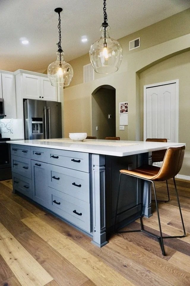 Modern kitchen with a large gray island, white countertop, brown leather bar stools, pendant lights, stainless steel fridge, wood flooring, and neutral walls. A bowl is on the island and an arched doorway is in the background.