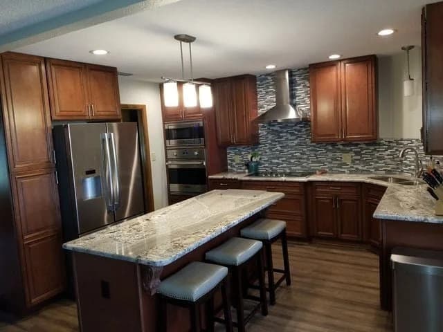 Modern kitchen with wooden cabinets, stainless steel appliances, a center island with three stools, granite countertops, and a mosaic tile backsplash under recessed lighting.