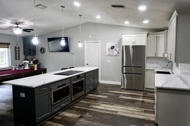 Modern kitchen with marble countertops, stainless steel appliances, and dark cabinets. An island with built-in ovens is centered. Adjacent dining area with a table and pendant lighting. Light gray walls and wood-look flooring complete the space.