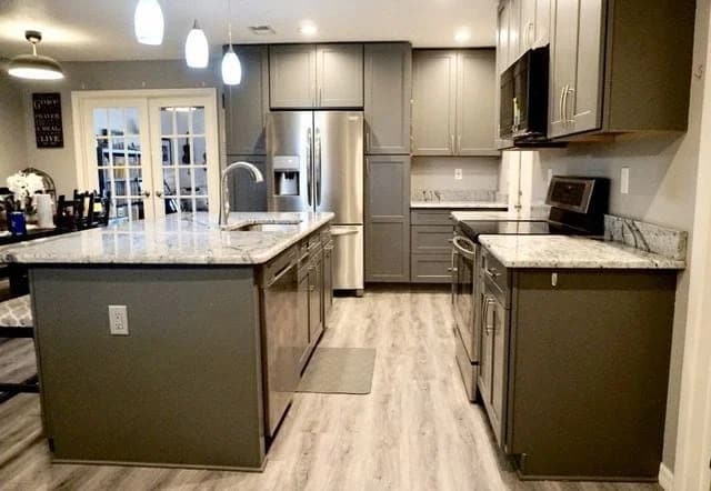 Modern kitchen with gray cabinets, marble countertops, a large island with a sink, stainless steel appliances, and wood-look flooring. Pendant lights hang above the island, and a dining area is visible in the background.