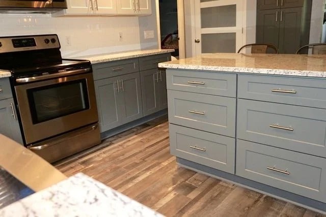 Modern kitchen with stainless steel oven, gray cabinets, granite countertops, and a large island. The floor has a wood-like finish and white upper cabinets are seen in the background.