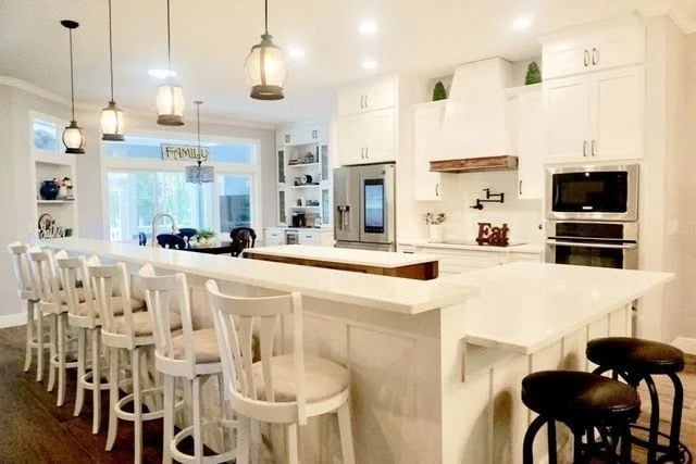 Bright, modern kitchen with white cabinets, a large island with white bar stools, pendant lights, built-in appliances, and a dining area in the background. The space is tidy and well-lit with natural and overhead lighting.