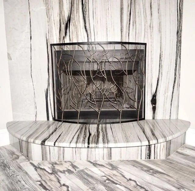 A modern fireplace with a black metal screen featuring a branch pattern, set against a wall, hearth, and floor with white and black marble-like streaks creating a bold, striped effect.