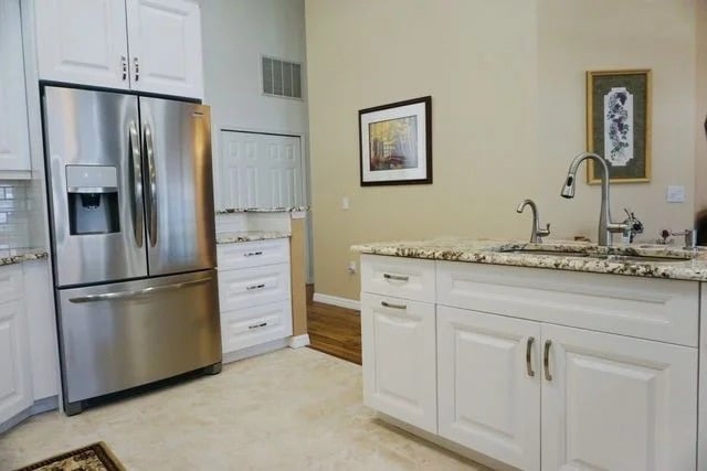 A modern kitchen with white cabinets, granite countertops, a stainless steel refrigerator, and a double sink. Artwork is hung on light-colored walls, and hardwood flooring is visible in the background.