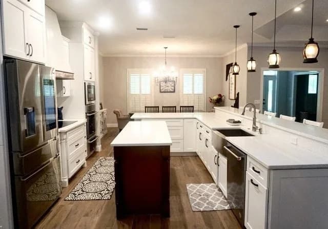 Modern kitchen with white cabinets, stainless steel appliances, a large central island, pendant lights, wooden floor, and an adjacent dining area with a table and chairs near a window with shutters.
