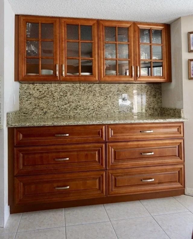 A kitchen area with wooden cabinets, glass-front upper cabinets displaying dishes, a granite countertop, matching granite backsplash, and tiled floor. The lower cabinets have large drawers with silver handles.
