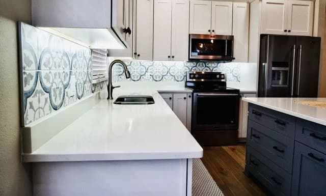 Modern kitchen with white cabinets, patterned blue and white tile backsplash, stainless steel appliances, a built-in microwave, and a large island with dark drawers and a white countertop.