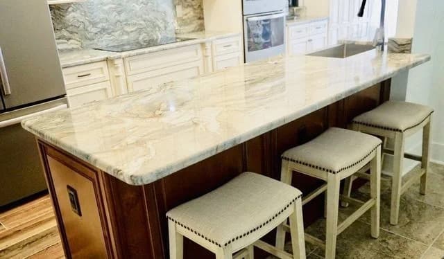 A kitchen with a long marble island countertop, three upholstered stools with nailhead trim, white cabinetry, stainless steel appliances, and a marble backsplash.