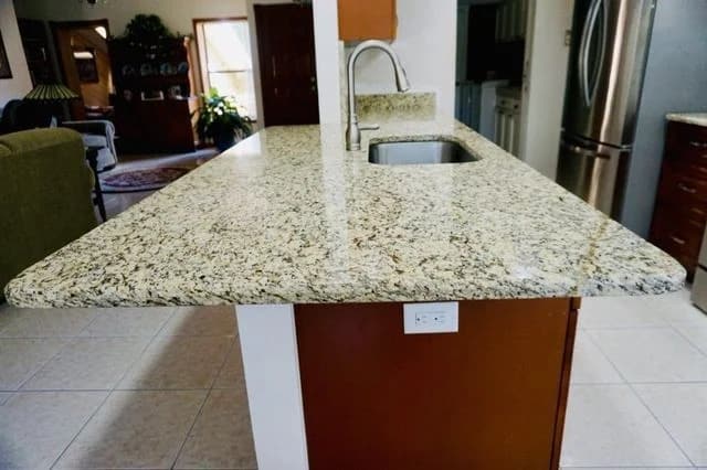 Granite kitchen countertop with a built-in sink and modern faucet, brown base cabinet, and electrical outlet. Stainless steel refrigerator and tiled floor are visible in the background.
