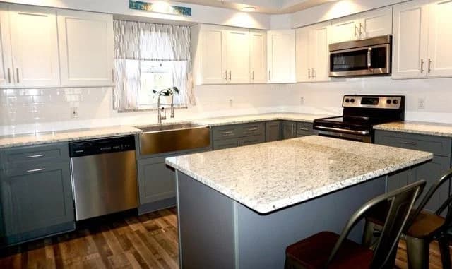 A modern kitchen with white upper cabinets, gray lower cabinets, a large granite island, stainless steel appliances, a farmhouse sink under a window, and wood-style flooring.