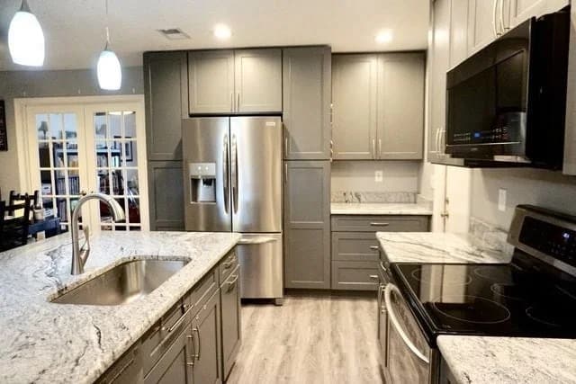 Modern kitchen with gray cabinets, stainless steel appliances, a marble countertop island with a sink, electric stove, refrigerator, and microwave. The flooring is light wood, and pendant lights hang over the island.