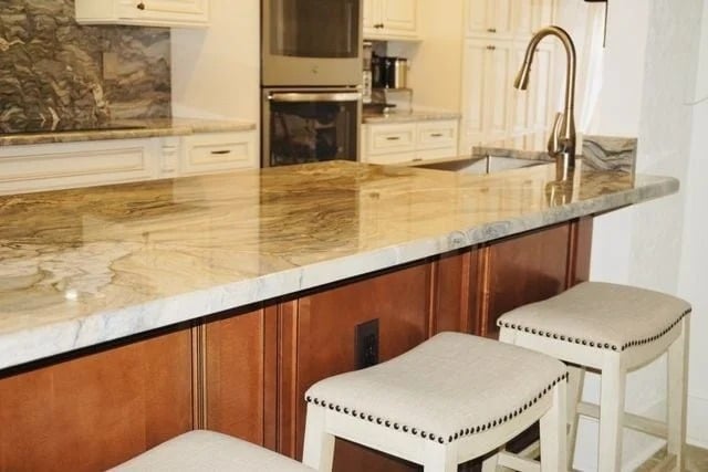 A modern kitchen with a marble countertop, stainless steel sink and faucet, wooden cabinets, and three upholstered bar stools with nailhead trim. White cabinetry and an oven are visible in the background.