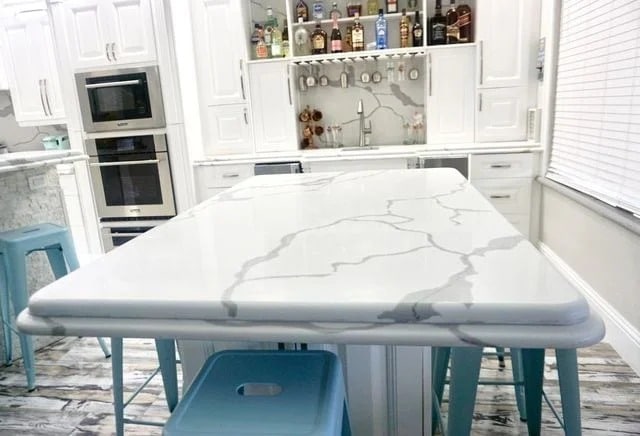 A kitchen with white cabinets, a marble island countertop with gray veins, light blue stools, and shelves holding bottles and cups above a small sink area. Light streams in from a window with blinds on the right.