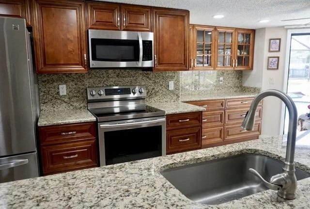 Modern kitchen with brown wooden cabinets, stainless steel appliances including an oven and microwave, granite countertops, a sink with a faucet, and glass-front upper cabinets.