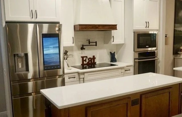 Modern kitchen with white cabinets, stainless steel appliances, a refrigerator with a touch screen, a stove with a white hood, a microwave, and a small island with a wooden base and white countertop.