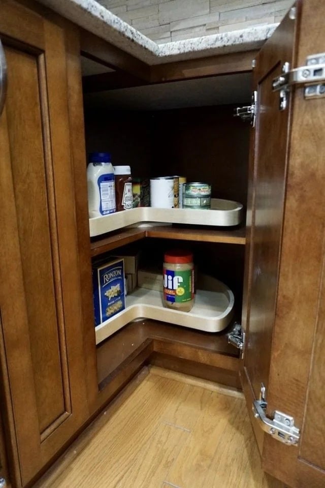 A wooden kitchen cabinet with open doors reveals two pull-out, kidney-shaped shelves containing pantry items like peanut butter, pasta, canned goods, and spices. The shelves are partially extended and organized.