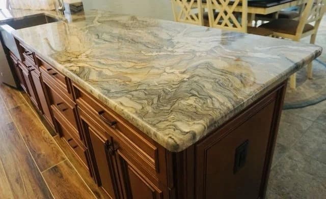 A kitchen island with a polished, multicolored marble countertop featuring wavy patterns, set atop dark wooden cabinets. The island stands on a wood floor, with chairs and a dining table visible in the background.